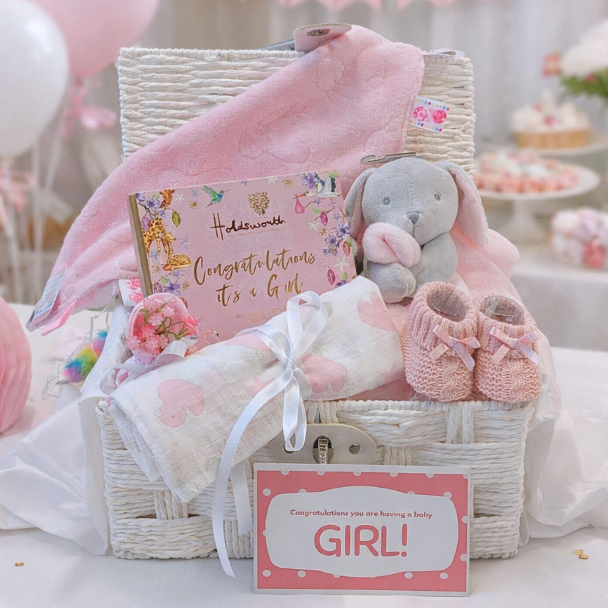 A pink baby girl gender reveal hamper basket with gifts including 'its a girl' chocolates, soft toy and gift card.