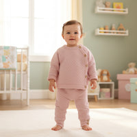 Baby girl wearing pink luxury knitted baby clothes set with honeycomb knit top and matching trousers in nursery.