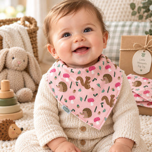 Baby wearing a pink bandana bib with squirrels, hedgehogs, flowers and green leaves.
