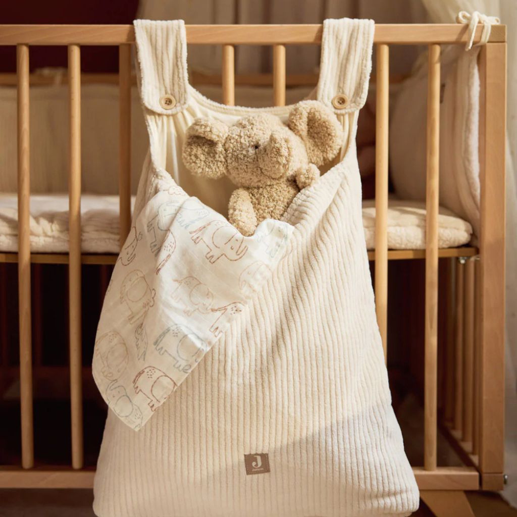 Beige baby bag with elephant teddy plush toy hanging on a wooden crib.
