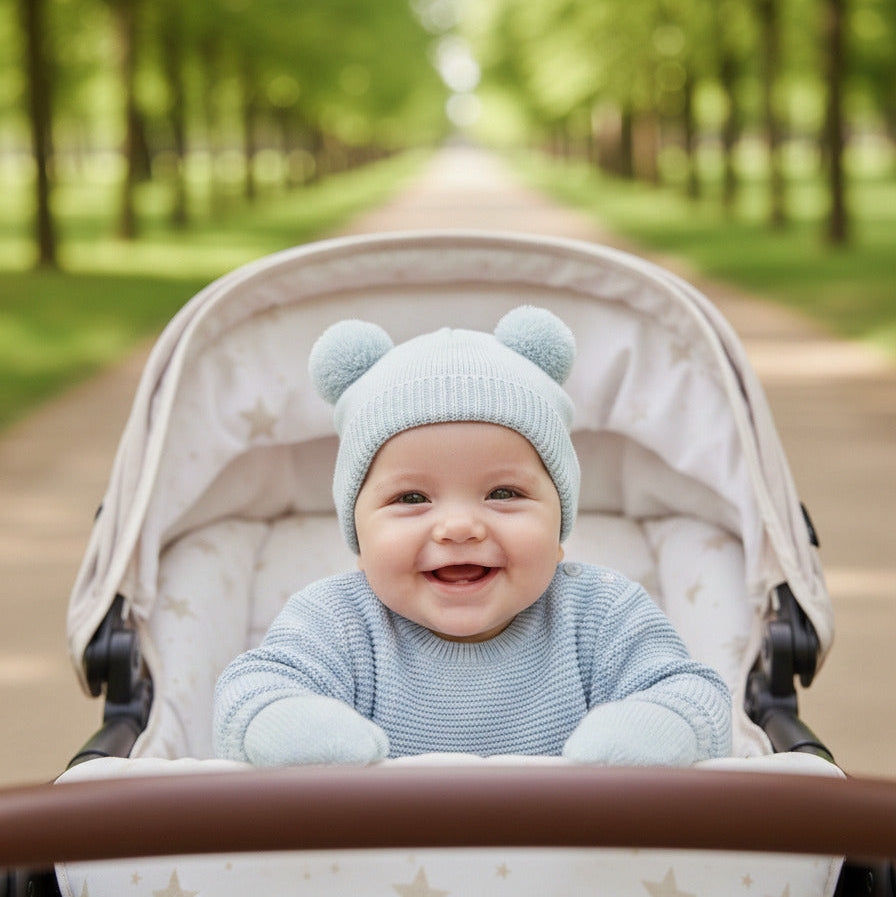 Baby in a stroller wearing a sage green knit hat with pom pom bear ears