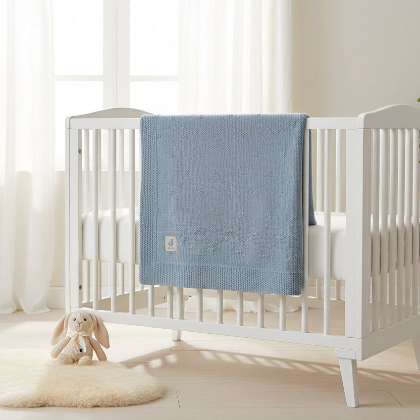 A sea green textured baby blanket draped over a white wooden crib in a bright, softly lit nursery with sheer curtains, a plush bunny toy, and a cream rug.