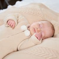 Baby wearing a taupe knitted jacket with ivory pom poms, shown close up while lying on a knitted blanket.