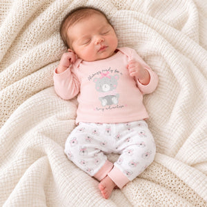 Baby wearing pink bear top and white bear-print trousers, with two wrist rattles beside her on a cream blanket.