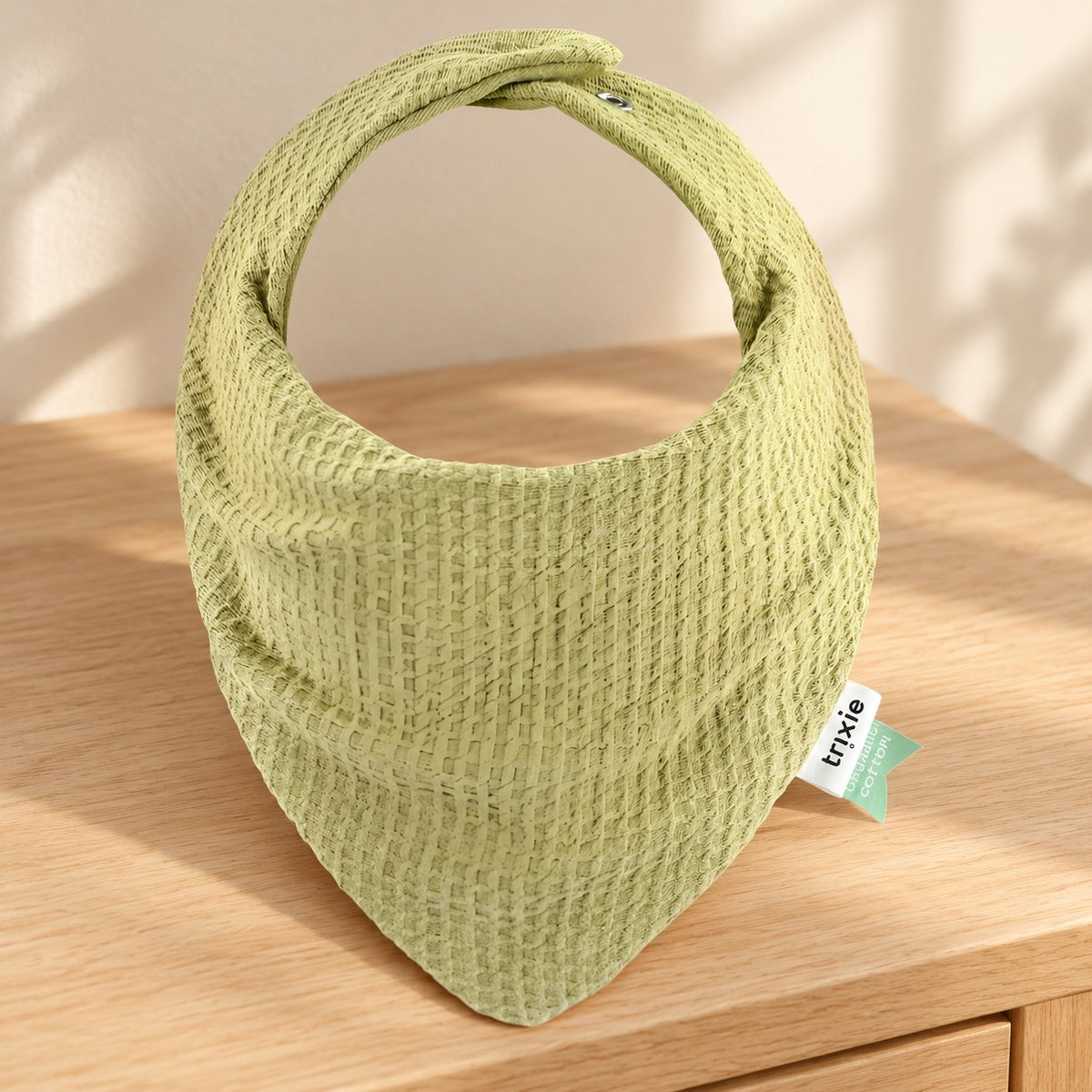 Lemongrass green textured bandana bib resting on a wooden dresser beside a softly lit cream wall.