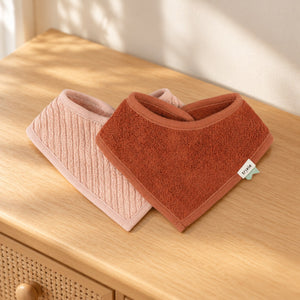 Pale pink and rust Hush Rose bandana bibs overlapping on a wooden dresser in soft daylight.