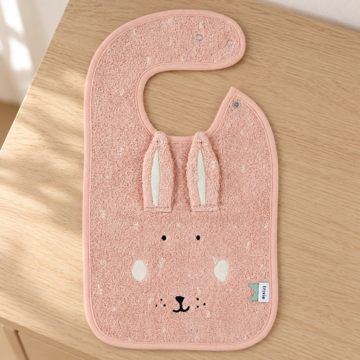 Pink Mrs Rabbit bib with long fabric ears and a rabbit face laid flat on a sunlit wooden dresser.