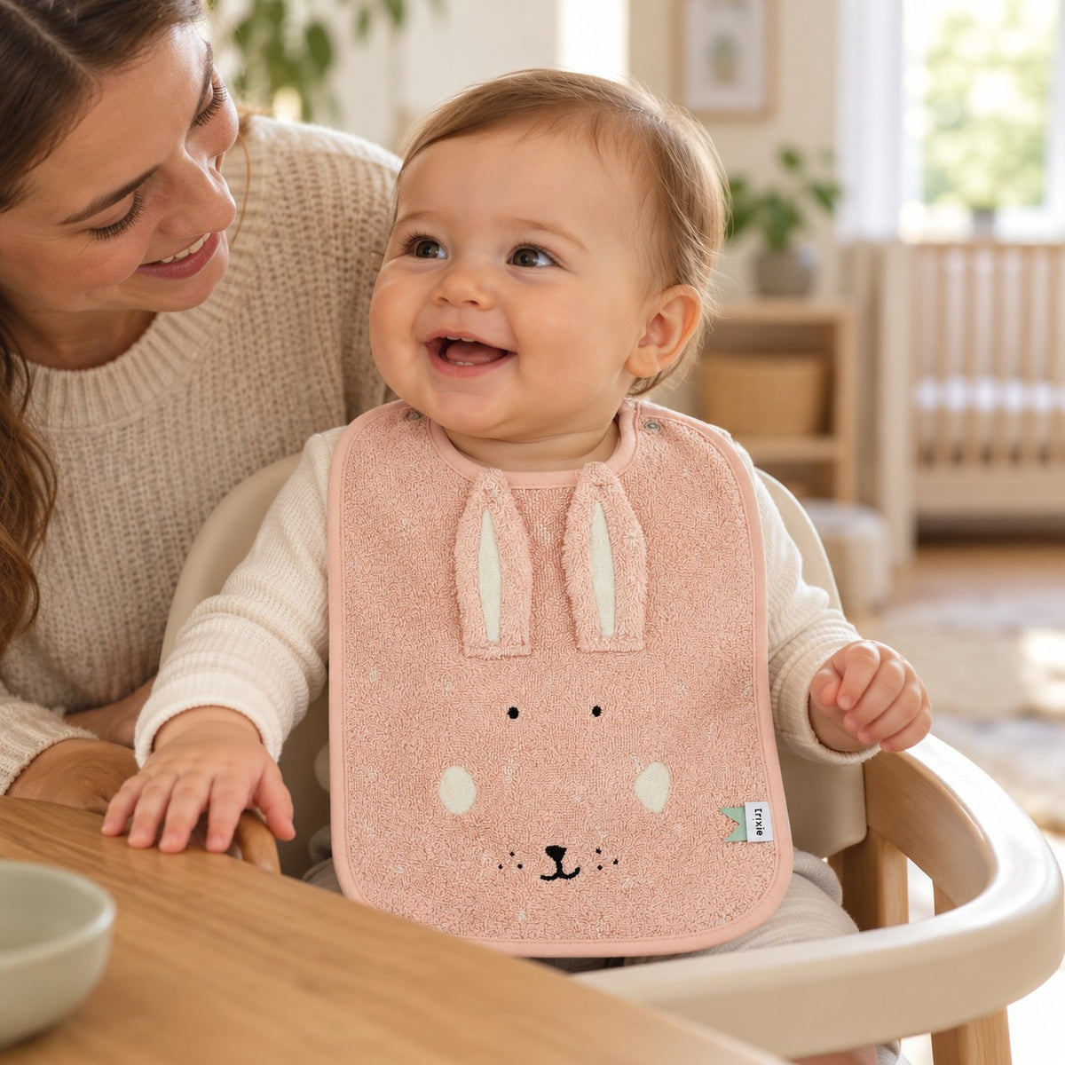 Baby wearing the pink Trixie Mrs Rabbit bib in a highchair beside a parent.