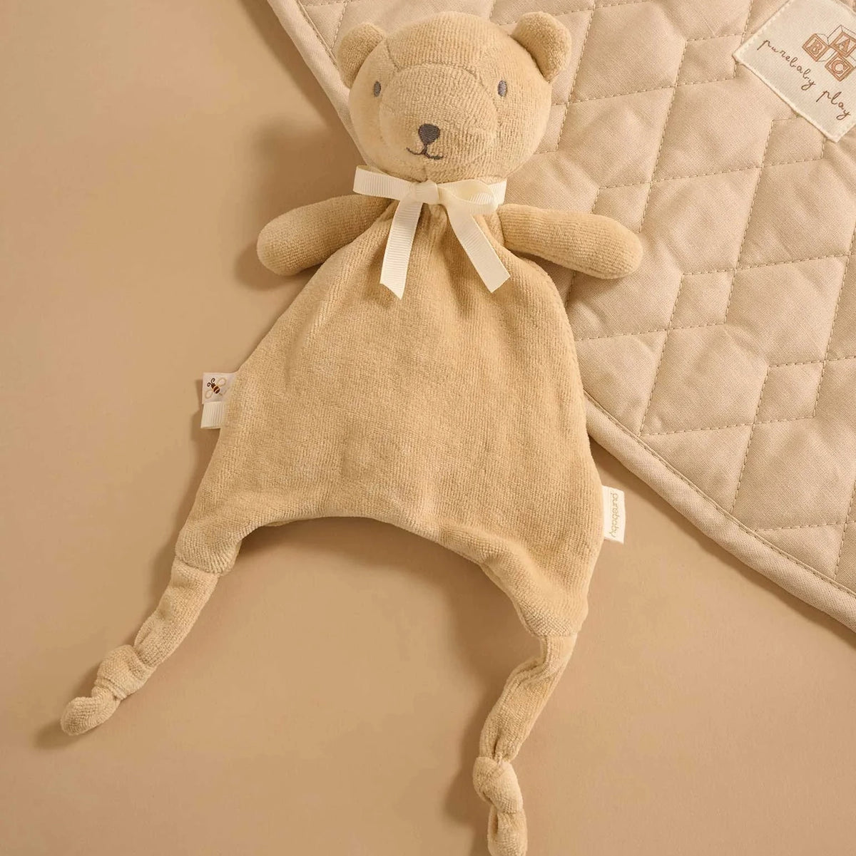 Wheat bear baby comforter laid flat, showing soft velour body, ribbon bow and knotted feet