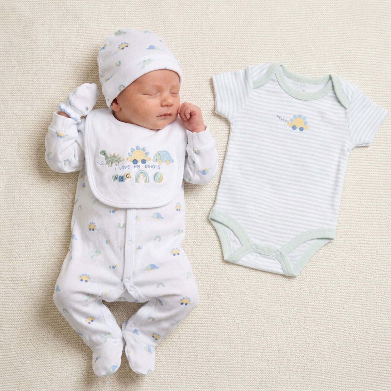 White baby clothes set with a dinosaur print. This 5 piece outfit includes sleepsuit, hat. mittens and bib worn by a baby with the bodysuit shown on the right.