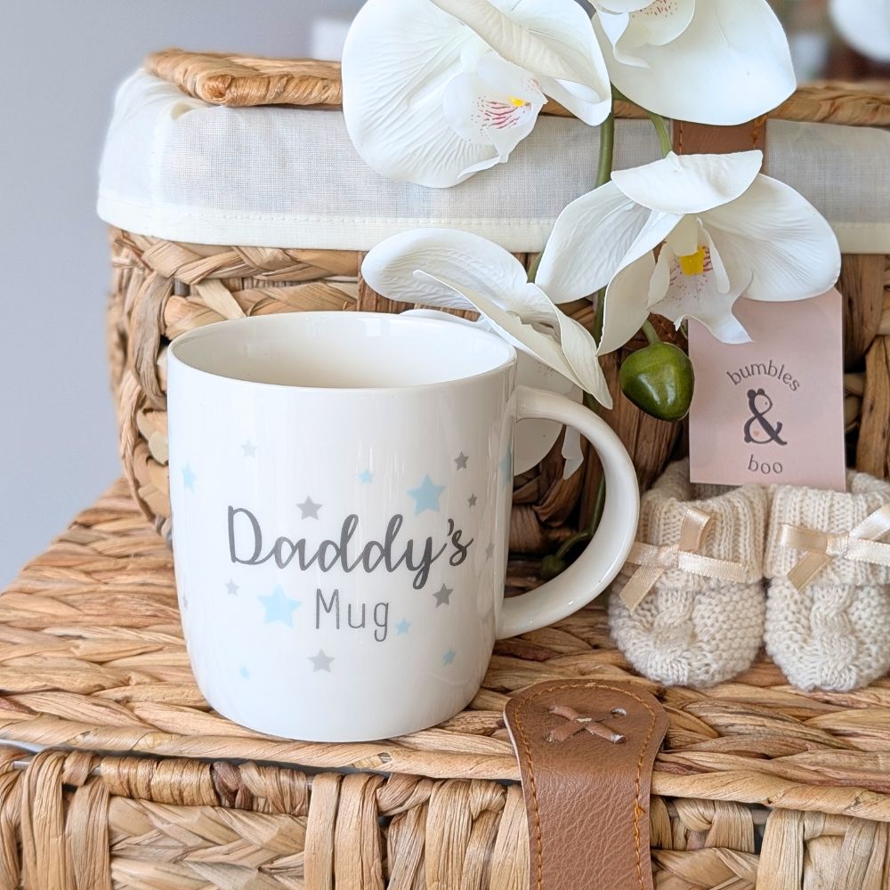 White “Daddy’s Mug” with blue and grey stars, styled with baby booties and a wicker basket.