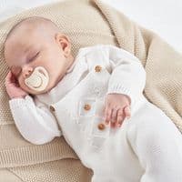 Close-up of a baby wearing white knitted dungarees and a collared jumper with wooden strap buttons.