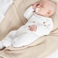 Baby lying on a knitted blanket in the white dungaree and jumper set, showing the footed legs.