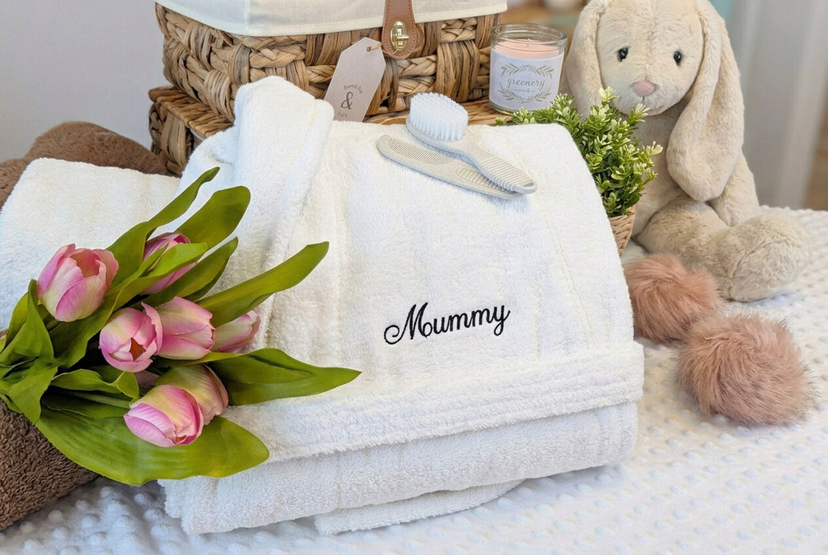 Folded white Mummy dressing gown with silver embroidery, displayed beside pink flowers and a gift basket.