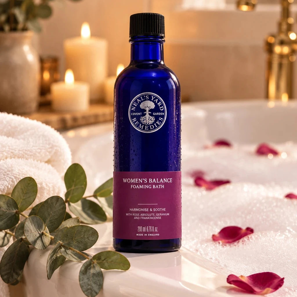 Women's Balance Foaming Bath 200 ml blue bottle beside a bubble bath with rose petals, candles and a white towel.