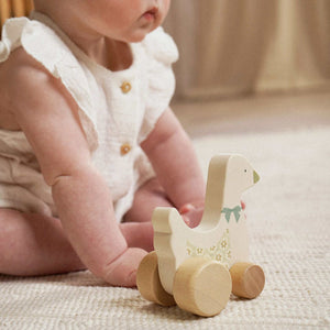 Child seated beside a cream wooden goose toy with floral detail and natural wood wheels.