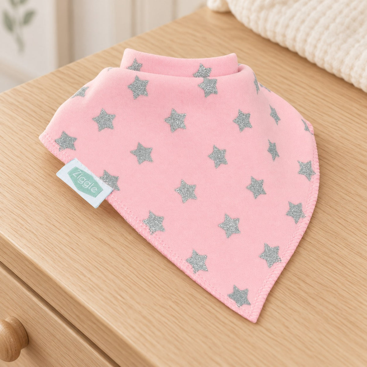 Pink Ziggle bandana bib with silver glitter stars displayed on a wooden nursery dresser.