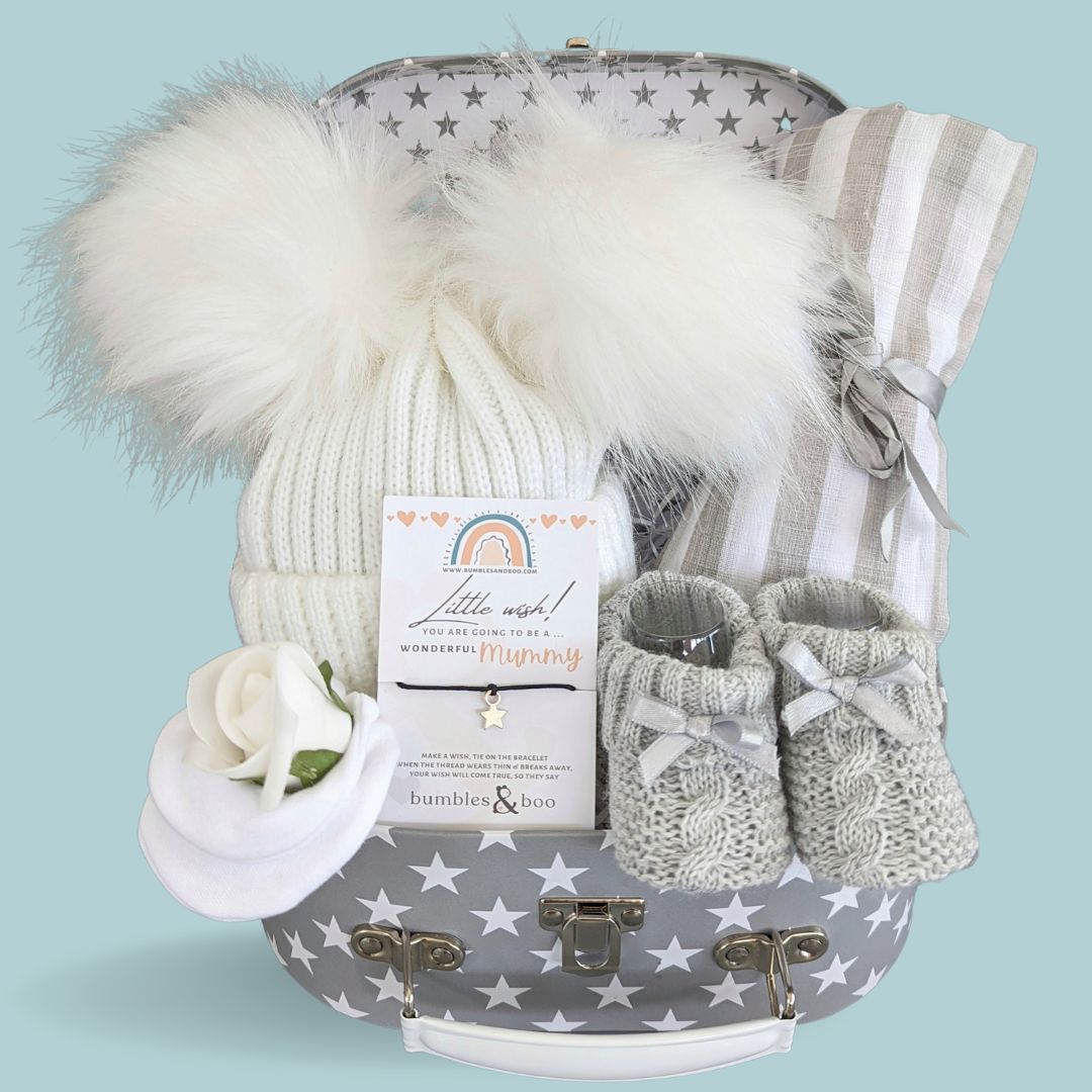 Baby shower hamper with gifts including white double pom pom hat, knit baby booties, mittens and grey striped muslin.