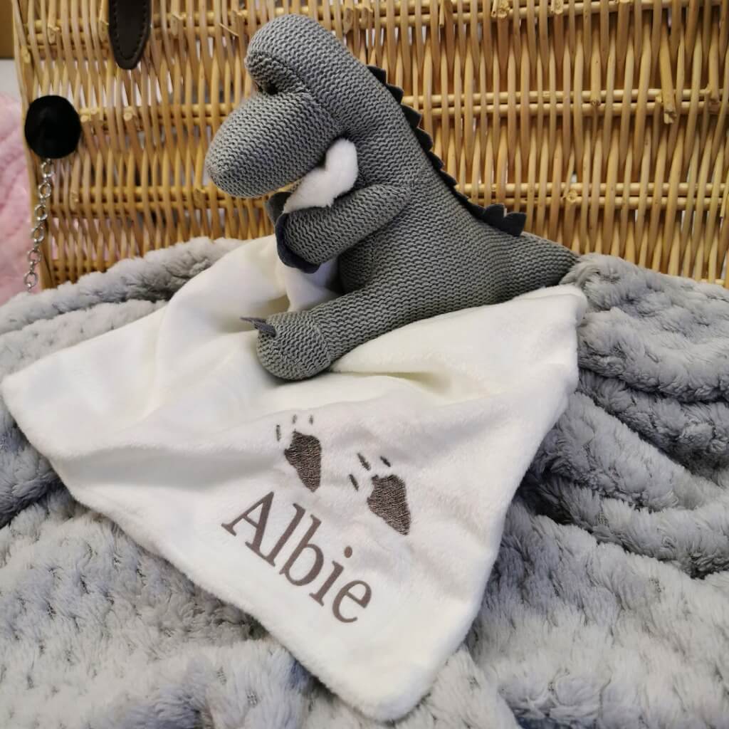 Personalised grey dinosaur baby comforter holding a white blanket with the baby&#39;s name.
