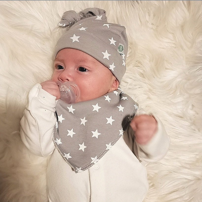 Baby wearing a grey bandana bib with white stars over white baby clothes, with a matching hat.